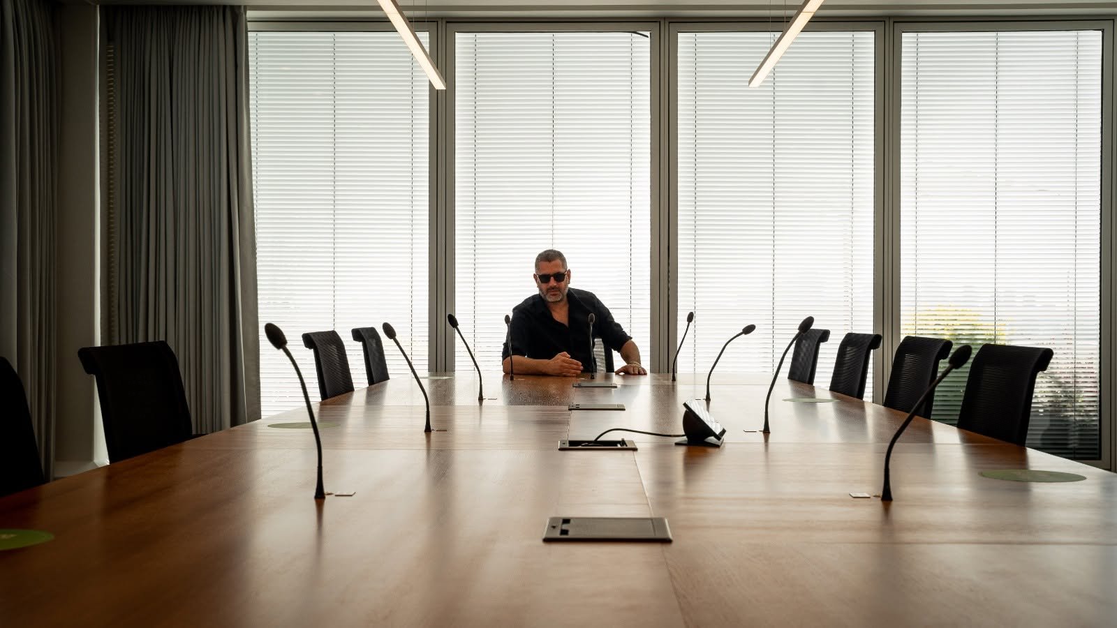 David seated alone at the head of a long boardroom table, black shirt and sunglasses