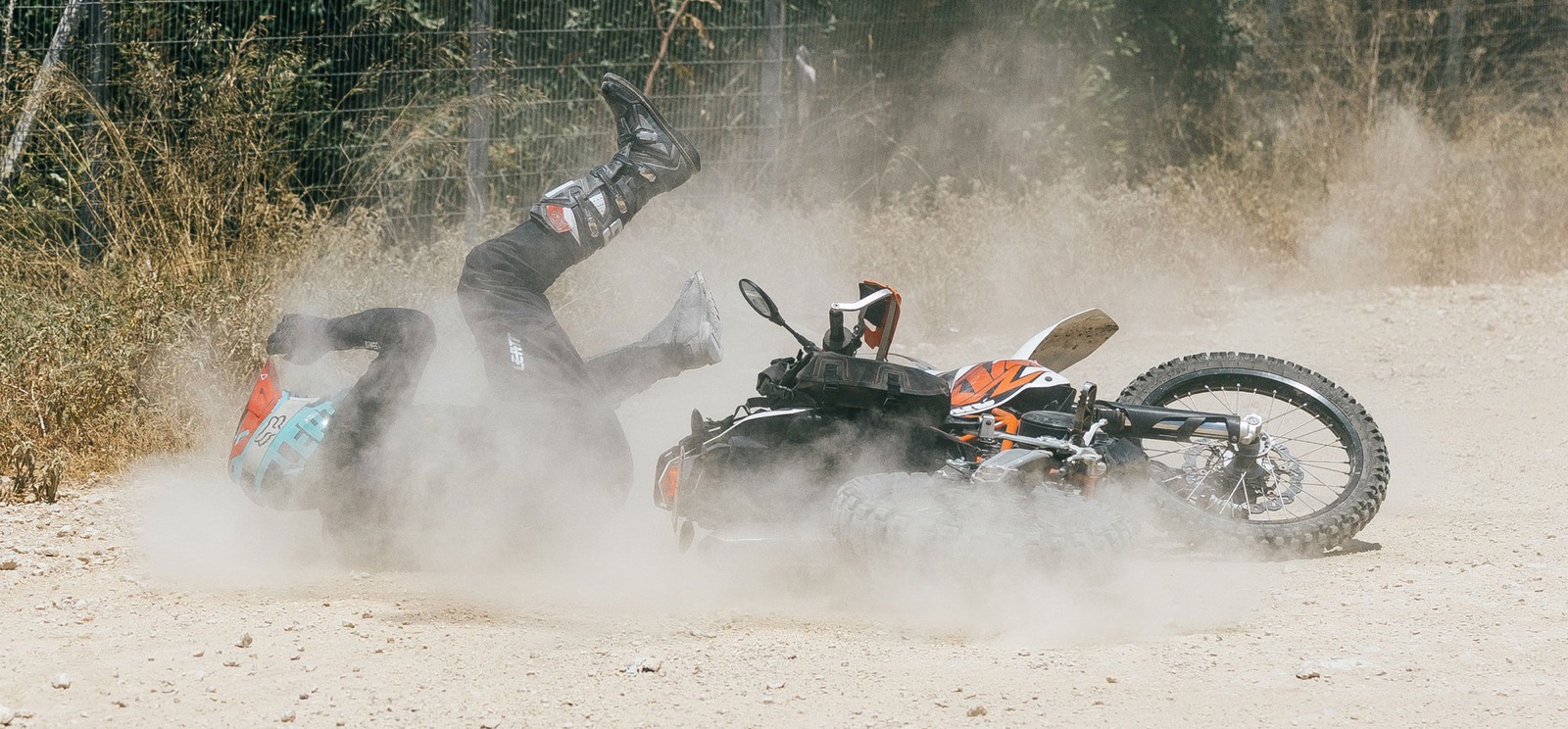 Rider tumbling off a KTM adventure bike in a cloud of dust on a gravel road