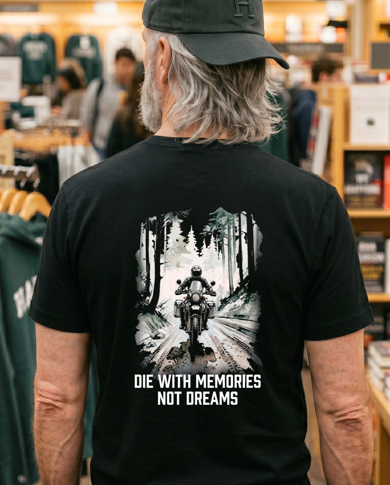 Gray-haired man seen from behind in a busy bookstore, wearing the black Die With Memories Not Dreams tee with its forest trail back print