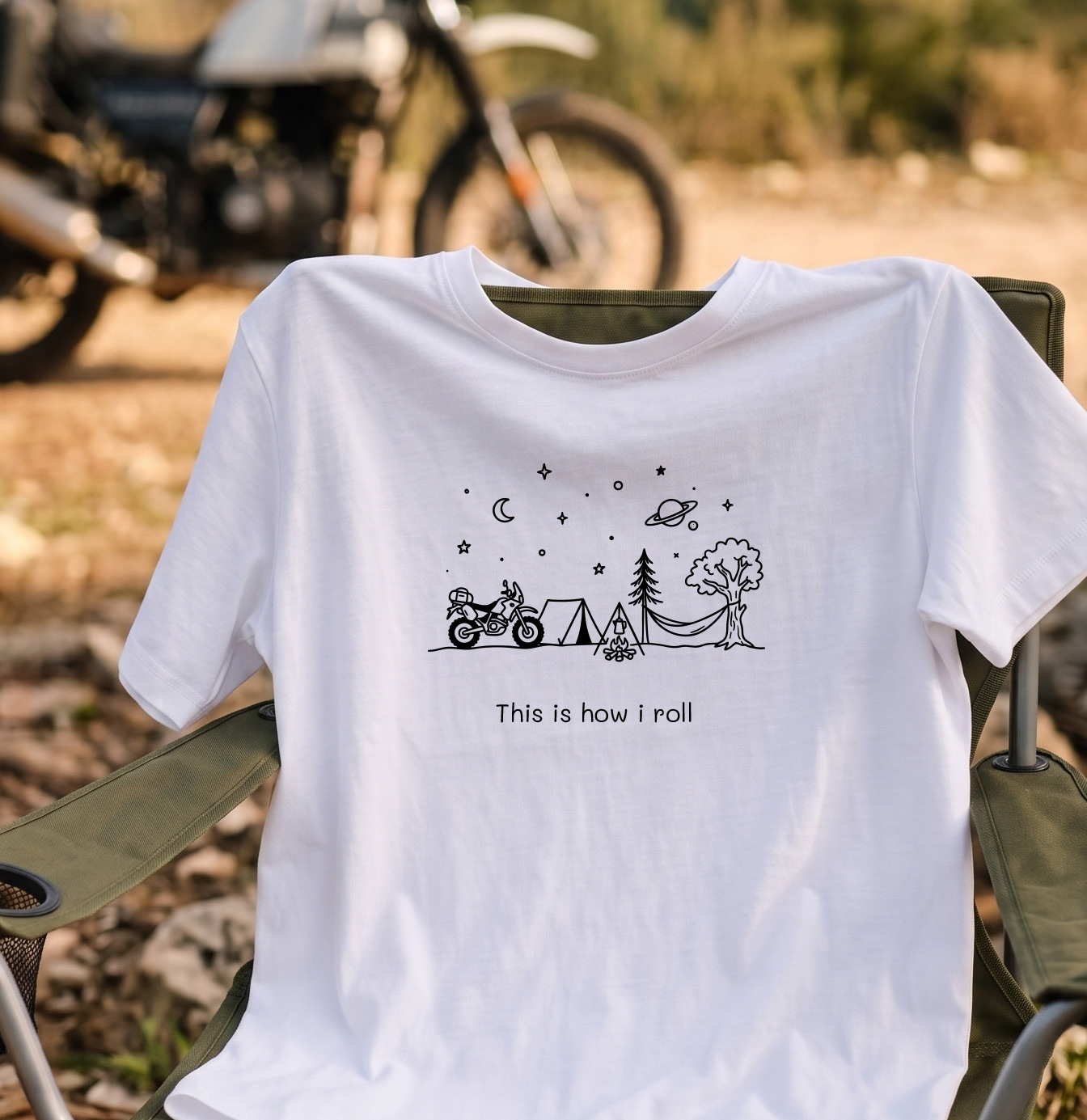 White tee with a line drawing of a campsite, tent, campfire and parked adventure bike reading This is how i roll, draped over a camp chair with a motorcycle blurred behind